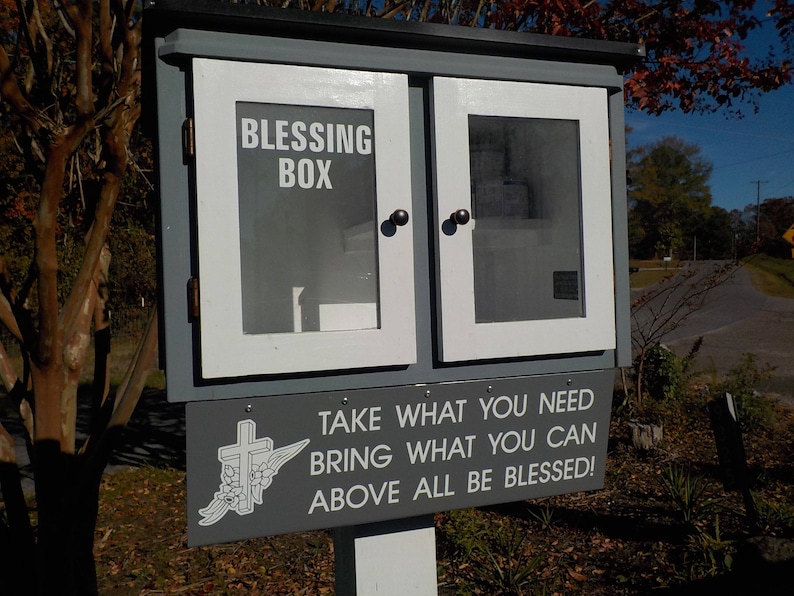 May include: A wooden box with a white door and a sign that reads "Blessing Box Take What You Need Bring What You Can Above All Be Blessed!"