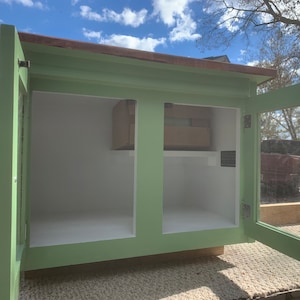 May include: A green wooden cabinet with two doors, one with a glass pane. The doors are open, revealing an empty interior with a shelf. The cabinet has a brown wooden top.