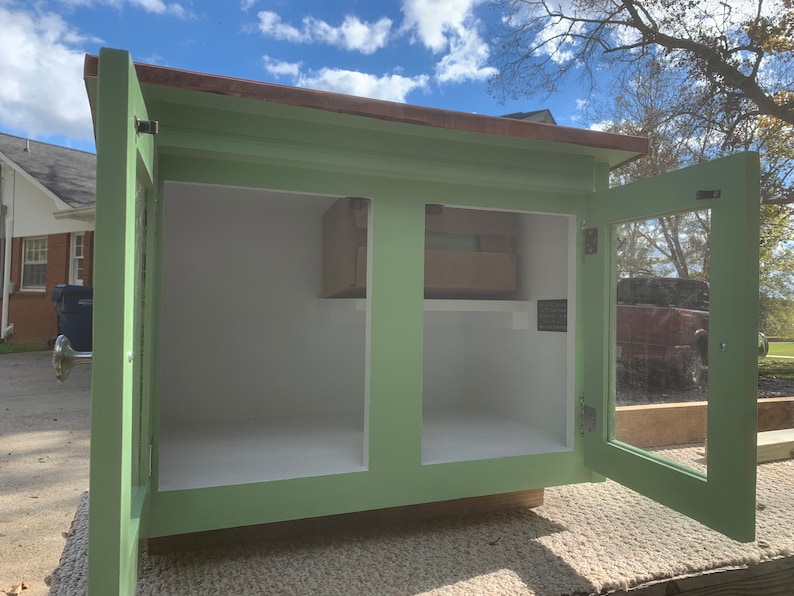 May include: A green wooden cabinet with two doors, one with a glass pane. The doors are open, revealing an empty interior with a shelf. The cabinet has a brown wooden top.