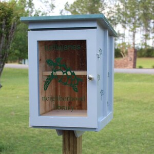 May include: A light blue wooden neighborhood library with a green roof, mounted on a wooden post. The front door has a clear window with a green turtle graphic and the words "Tortolanes neighborhood library".