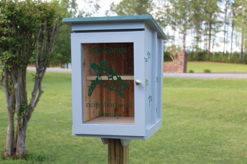 May include: A light blue wooden neighborhood library with a green roof, mounted on a wooden post. The front door has a clear window with a green turtle graphic and the words "Tortolanes neighborhood library".