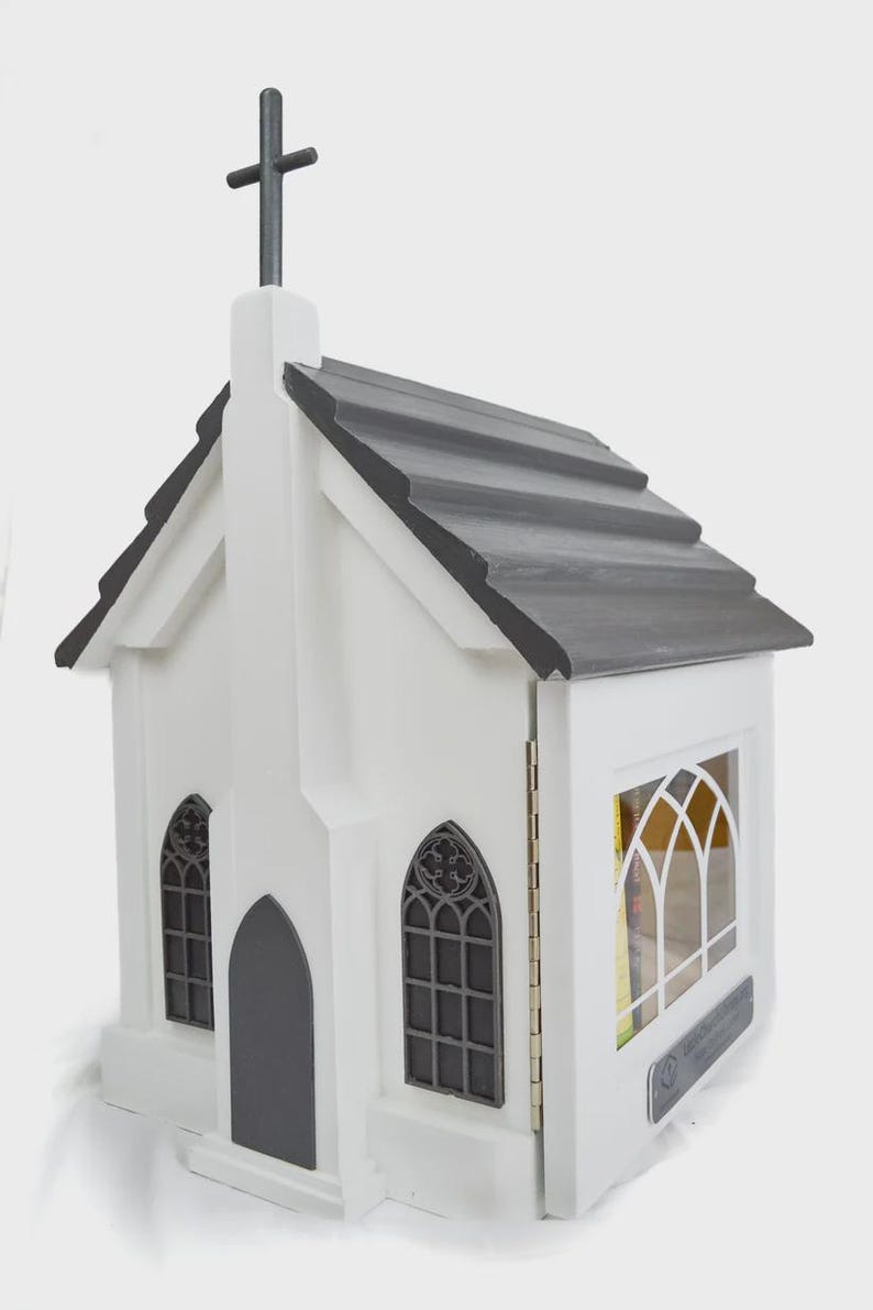 May include: A white wooden church-shaped box with a gray roof and a cross on top. The box has arched windows and a dark gray door. The side of the box opens to reveal a shelf. The box is designed to hold books or other small items.