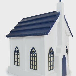 May include: A white wooden church model with a navy blue roof and cross. The model has arched windows with dark blue trim and a matching door. The church is a miniature replica of a traditional church building.