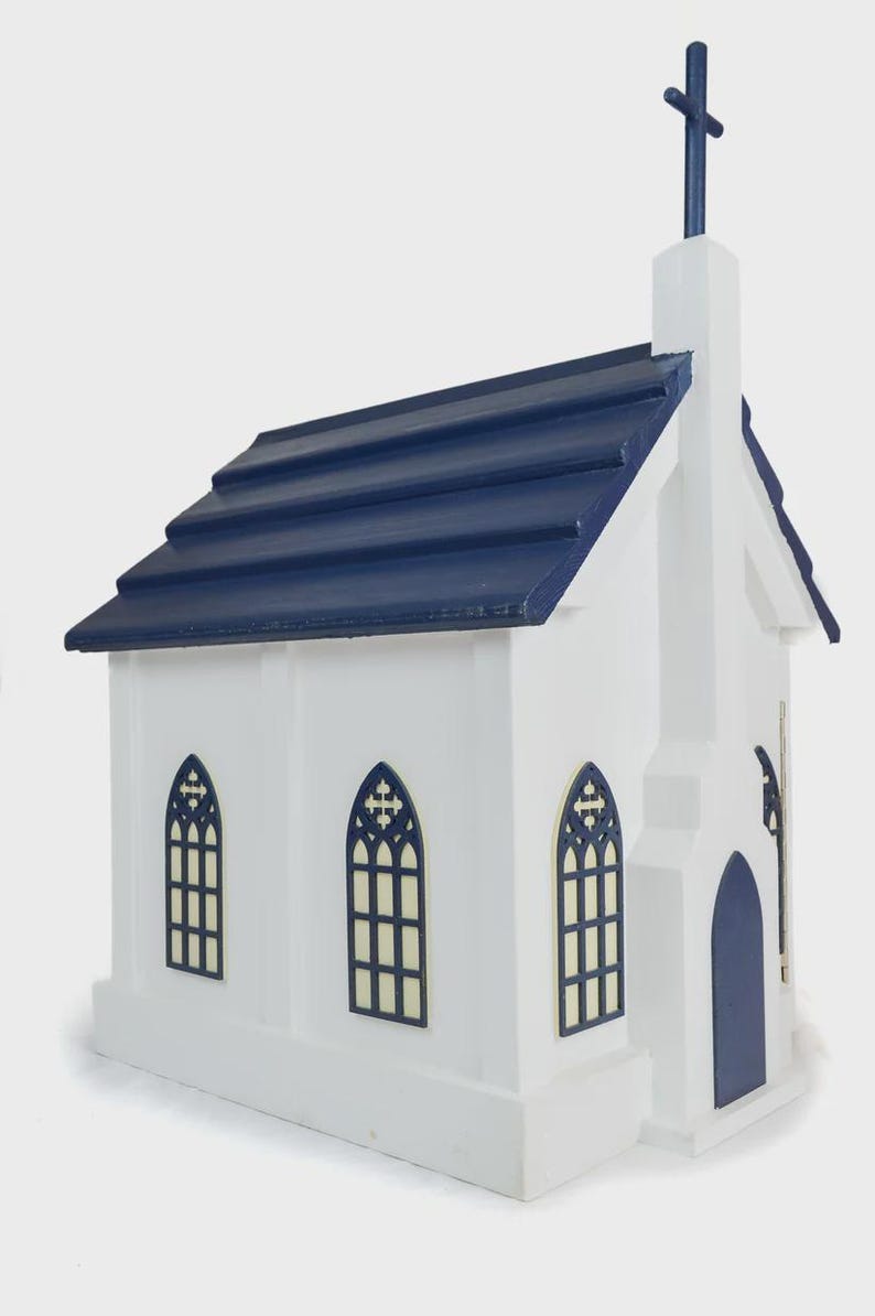 May include: A white wooden church model with a navy blue roof and cross. The model has arched windows with dark blue trim and a matching door. The church is a miniature replica of a traditional church building.