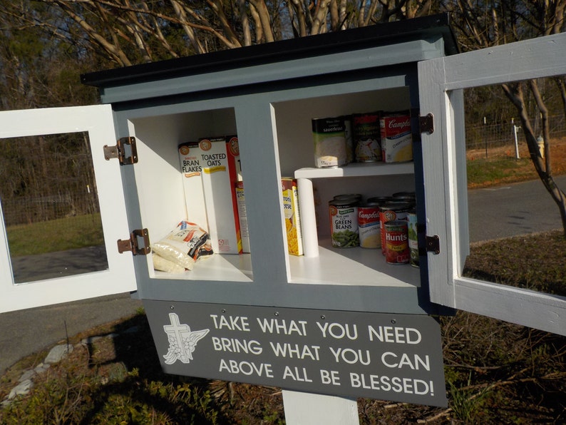 May include: A gray wooden cabinet with glass doors open, revealing shelves stocked with canned goods and boxed food. The cabinet has a sign that reads "TAKE WHAT YOU NEED BRING WHAT YOU CAN ABOVE ALL BE BLESSED!"