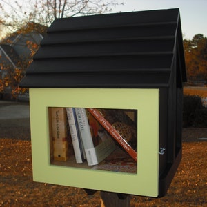 May include: A small, black and yellow wooden free library with a pitched roof. The library has a window displaying several books. The background shows a residential area with trees and a house.