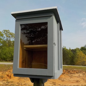May include: A gray wooden Little Free Library with a clear glass door and a small wooden shelf inside. The library is mounted on a wooden post and has a sloped roof. The background includes trees and a blue sky.