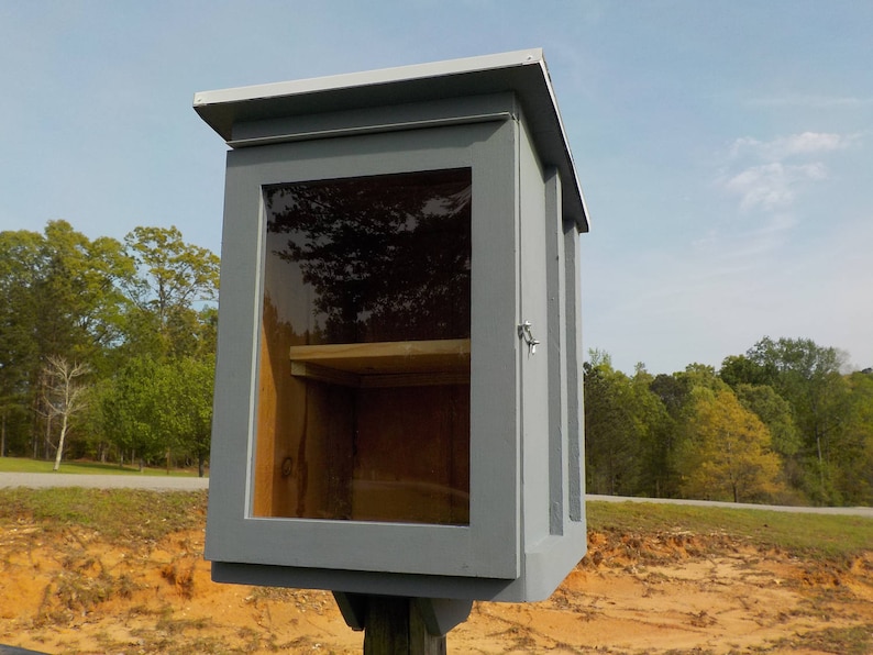 May include: A gray wooden Little Free Library with a clear glass door and a small wooden shelf inside. The library is mounted on a wooden post and has a sloped roof. The background includes trees and a blue sky.