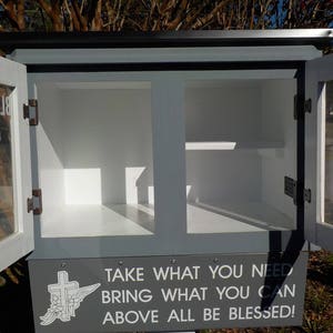 May include: A gray wooden box with glass doors and a white sign that reads "Blessing Box Take What You Need Bring What You Can Above All Be Blessed!" The box is open and empty.