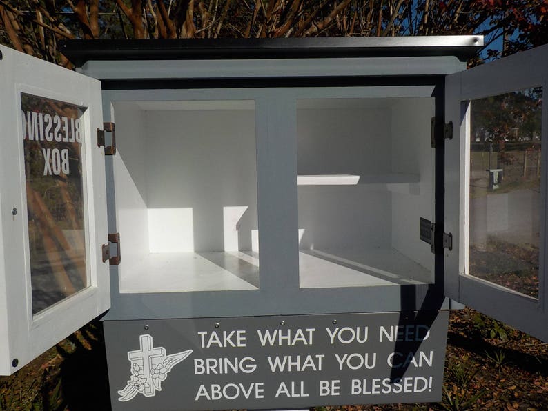 May include: A gray wooden box with glass doors and a white sign that reads "Blessing Box Take What You Need Bring What You Can Above All Be Blessed!" The box is open and empty.