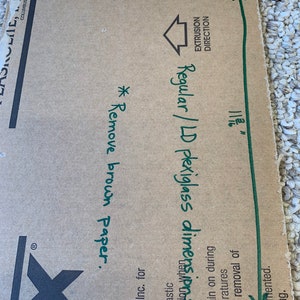 May include: A close-up of a brown cardboard box with handwritten instructions for removing brown paper before using regular or LD plexiglass. The box is labeled with the dimensions 7 3/4 inches by 11 1/2 inches. The text "EXTRUSION DIRECTION" is also visible on the box.
