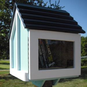 May include: A light blue wooden book exchange box with a black shingled roof. The box has a white frame around the opening and a glass window. The window shows several books with titles including "Dead or Alive" by Tom Clancy and "The Assassin" by Clive Cussler.