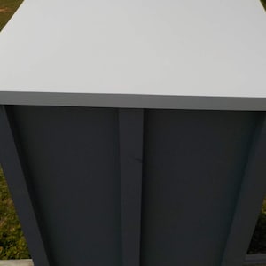 May include: A rectangular, light gray wooden box with a white top. The sides are a darker gray with vertical and horizontal trim. The box is outdoors, with grass and a wooden fence visible in the background.