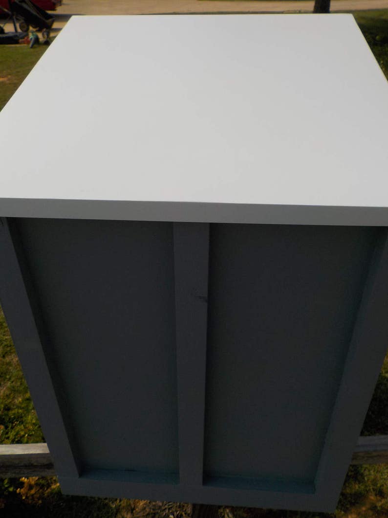 May include: A rectangular, light gray wooden box with a white top. The sides are a darker gray with vertical and horizontal trim. The box is outdoors, with grass and a wooden fence visible in the background.