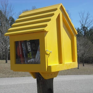 May include: A bright yellow wooden book exchange box with a sloped roof and a glass door. The door is open and several books are visible inside. The box is mounted on a wooden post.
