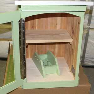May include: A small, open-door wooden cabinet painted in a soft green. The interior features a light wood shelf and a ramp-like structure. The cabinet has a white top and is constructed with visible hinges.