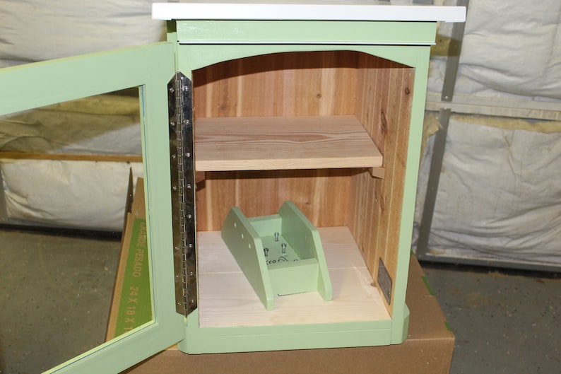 May include: A small, open-door wooden cabinet painted in a soft green. The interior features a light wood shelf and a ramp-like structure. The cabinet has a white top and is constructed with visible hinges.