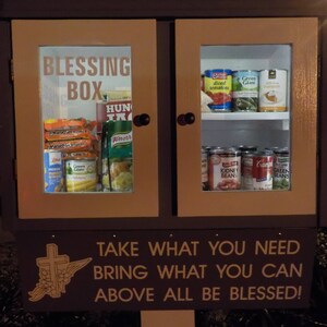 May include: A wooden cabinet with a glass door labeled "Blessing Box". The cabinet is filled with canned goods and other non-perishable food items. A sign below the cabinet reads "Take what you need, bring what you can, above all be blessed!"