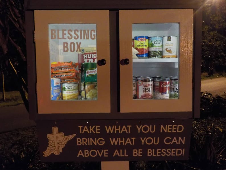 May include: A wooden cabinet with a glass door labeled "Blessing Box". The cabinet is filled with canned goods and other non-perishable food items. A sign below the cabinet reads "Take what you need, bring what you can, above all be blessed!"
