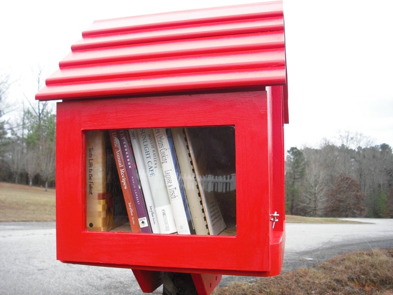 May include: A red wooden free library with a pitched roof and a window displaying a variety of books. The books include titles like "Lagasse & Tirsch", "Sunlight Cafe", "Home Annual Recipes", and "Tante Life to the Fullest".
