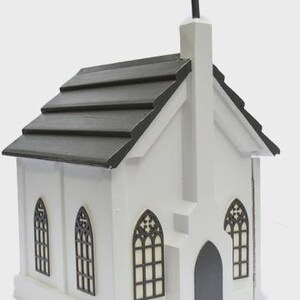 May include: A white miniature church with a black roof and cross. The church features arched windows with decorative black frames and a dark gray door. The overall design is a simple, yet elegant, representation of a church.