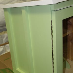 May include: A light green cabinet with a white top. The cabinet has a rectangular shape with a door on the right side. The door is attached with a long silver hinge. The cabinet is sitting on a cardboard box.
