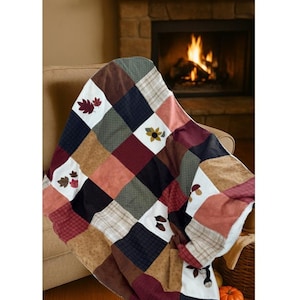 Puede incluir: Una manta de patchwork con cuadrados en tonos tierra, incluyendo marrón, rojo y beige. La manta presenta aplicaciones de hojas y flores. El texto "Manta 'Bosque dorado'" es visible.