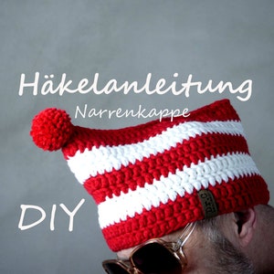 Könnte beinhalten: Ein rot-weiß gestreifter Häkelhut mit rotem Bommel. Der Hut ist mit "Häkelanleitung Narrenkappe DIY" beschriftet. Der Hut ist handgefertigt und scheint ein DIY-Projekt zu sein.