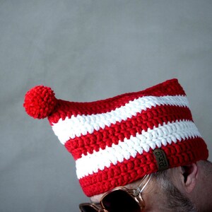 Può includere: Un cappello a uncinetto a righe rosse e bianche con un pon pon bianco in cima. Il cappello ha un'etichetta in pelle marrone con il testo "Original Handmade" su di essa.