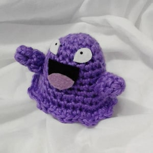 May include: A handmade purple crochet figure with white eyes and a black and purple mouth. The figure has a rounded body and outstretched arms, giving it a friendly appearance. The item is likely a toy or decorative item.