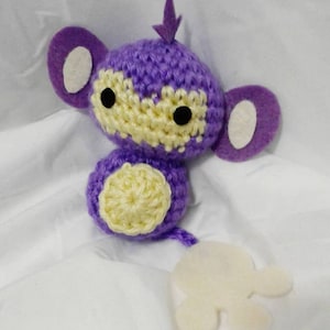 May include: A handmade crochet plush toy in the shape of a purple creature with a yellow face and belly. The toy has black eyes, large purple ears with white centers, and a small purple spike on its head. A white felt shape hangs from the bottom.