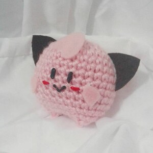 May include: A handmade, pink crocheted plush toy with black felt ears and a small pink horn. The toy has a smiling face with black eyes and a black mouth, and red cheeks. The plush is round and has small feet.