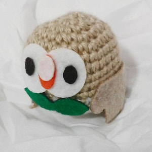 May include: A handmade owl-shaped crochet item. The owl features large white felt eyes with black pupils, a pink nose, and an orange beak. The body is crocheted in a light brown yarn, with felt wings and green felt leaves.