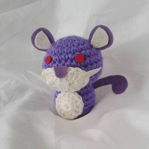 May include: A handmade crochet plush toy in the shape of a purple rodent with a white belly and face. The toy has red eyes, white whiskers, and a purple tail and ears. The plush is sitting on a white surface.