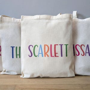 May include: Cream-coloured canvas tote bags with colourful text. The middle bag displays the name "SCARLETT" in rainbow colours. The other bags have partial names. The bags have a simple design with a handle at the top.
