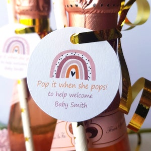 May include: Round white tag with a rainbow design in shades of pink, orange, and brown with black polka dots. The tag has the text "Pop it when she pops! to help welcome Baby Smith".