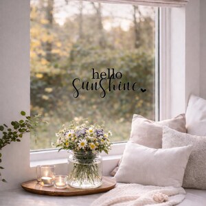 May include: A cozy window seat with a bouquet of daisies and other wildflowers in a glass vase. The scene includes lit candles on a wooden tray, soft pillows, and a knitted throw. The words "hello Sunshine" are written on the window.