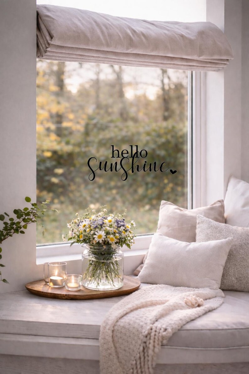 May include: A cozy window seat with a bouquet of daisies and other wildflowers in a glass vase. The scene includes lit candles on a wooden tray, soft pillows, and a knitted throw. The words "hello Sunshine" are written on the window.