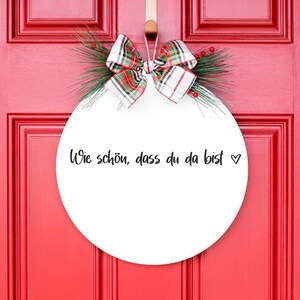 May include: White round door hanger with a red and green plaid bow and greenery. The hanger has the text "Wie sch&ouml;n, dass du da bist" written in black.