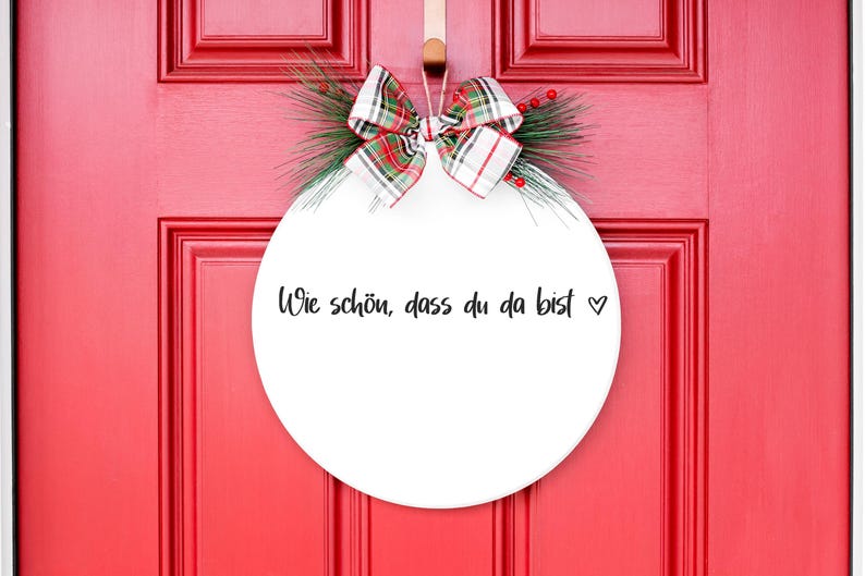 May include: White round door hanger with a red and green plaid bow and greenery. The hanger has the text "Wie sch&ouml;n, dass du da bist" written in black.