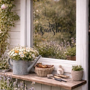 May include: A rustic outdoor scene featuring a window with the text "hello Sunshine". A galvanized metal watering can holds daisies. A wooden shelf displays a woven basket, a bird figurine, and potted plants. Roses and lavender add color.