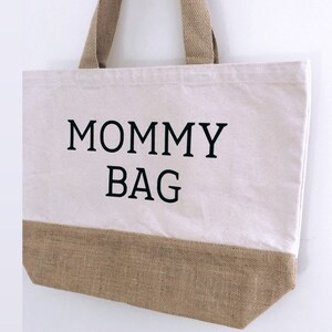 Puede incluir: Una bolsa de lona blanca con ribete de arpillera marr&oacute;n. La bolsa tiene texto negro que dice "MOMMY BAG".