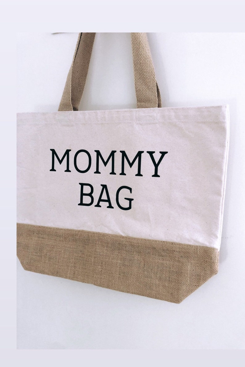 Puede incluir: Una bolsa de lona blanca con ribete de arpillera marr&oacute;n. La bolsa tiene texto negro que dice "MOMMY BAG".