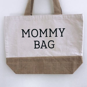 Puede incluir: Una bolsa de lona blanca con ribete y asas de yute marr&oacute;n. La bolsa tiene el texto "MOMMY BAG" impreso en negro.