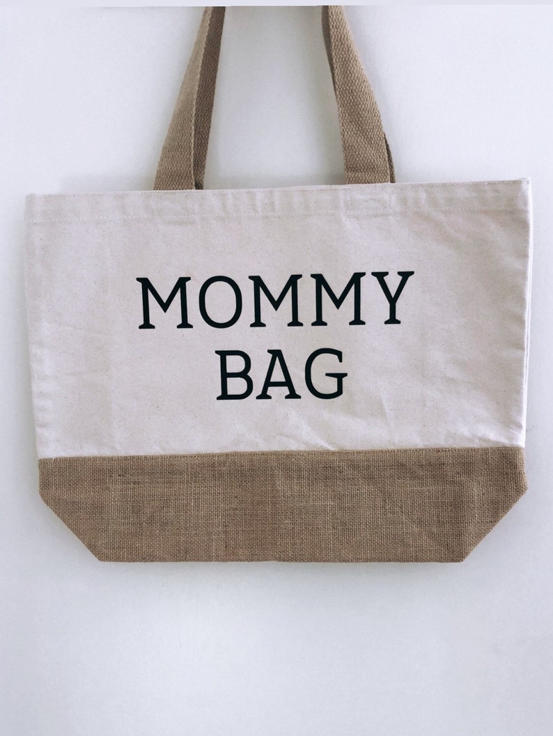 Puede incluir: Una bolsa de lona blanca con ribete y asas de yute marr&oacute;n. La bolsa tiene el texto "MOMMY BAG" impreso en negro.