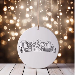 May include: A white, round Christmas ornament with a black line drawing of a village scene, including houses, trees, and a star-topped tree. The ornament hangs from a natural-colored string against a blurred background of warm lights.