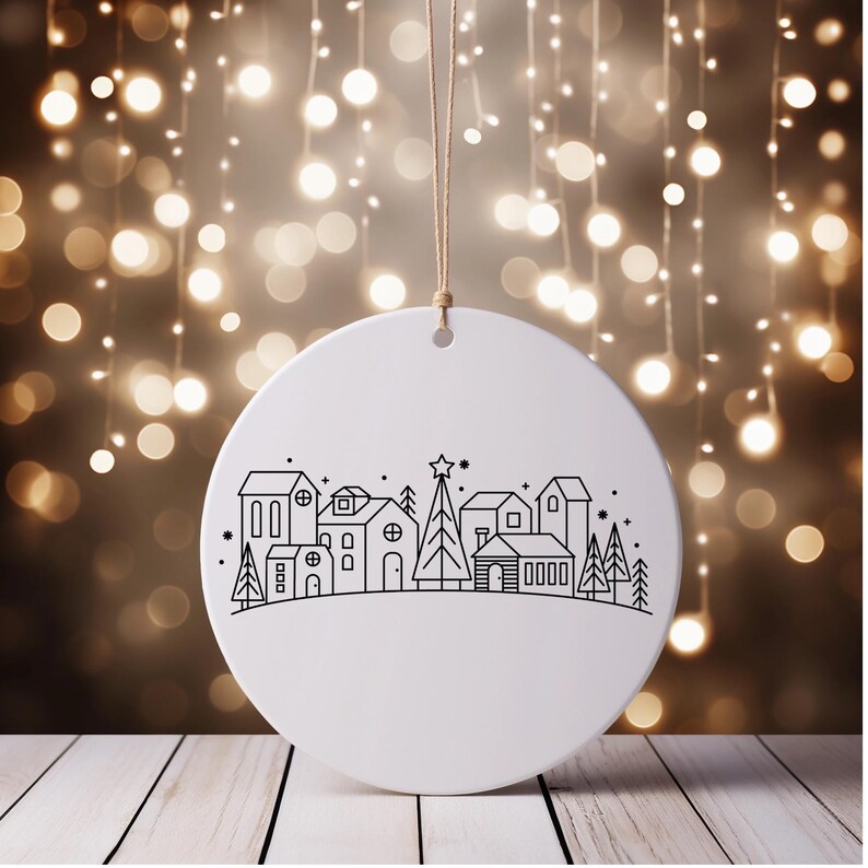 May include: A white, round Christmas ornament with a black line drawing of a village scene, including houses, trees, and a star-topped tree. The ornament hangs from a natural-colored string against a blurred background of warm lights.