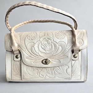 May include: Cream-colored leather handbag with a floral embossed design. The bag features a braided handle and a clasp closure. The front of the bag showcases a large rose surrounded by leaves, with additional floral details.