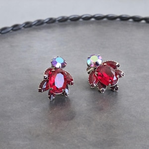 May include: A pair of vintage clip-on earrings. Each earring features a large, oval, ruby-red gemstone surrounded by smaller, dark red petals. A smaller, iridescent stone sits atop each earring. The earrings are set on a silver-toned metal.