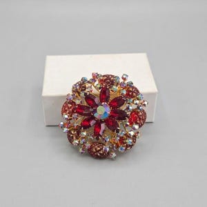 May include: A circular brooch featuring a central iridescent stone surrounded by deep red marquise-cut crystals. The outer edge is adorned with smaller, round, light-reflecting crystals set in a gold-tone metal frame with a floral design.